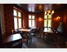 Gemütlicher Raum in einem Café oder Restaurant mit roten Tapeten im Retro-Stil, großen Fenstern, dunklen Holzstühlen und -tischen, sowie einer dekorativen Wandleuchte und einem Kronleuchter. Im Hintergrund ein ausgestelltes Kleid und Kerzen auf den Tischen für stimmungsvolle Beleuchtung.