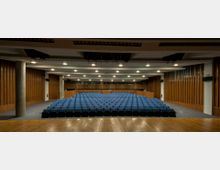 Ein moderner Hörsaal mit einer Holz-Bühne und mehreren Reihen blauer, gepolsterter Sitze. Die Wände sind mit vertikalen Holzpaneelen verkleidet, und die Decke ist mit eingebauten Leuchten und Akustikelementen gestaltet.