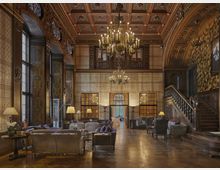 Großer, opulenter Innenraum eines historisch gestalteten Gebäudes mit verzierten Holzwänden und -decken. Zentral hängen große Kronleuchter, es gibt eine massive Holztreppe auf der rechten Seite und diverse Sitzgruppen mit Sofas und Sesseln, die den Raum im Stil eines eleganten Salons ergänzen.