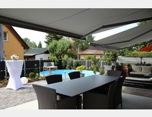 Eine moderne Terrasse mit Steinboden, möbliert mit einem dunklen Esstisch und Korbstühlen, im Vordergrund. Im Hintergrund sind ein kleiner runder Pool, Pflanzen in Töpfen, eine gepolsterte Sitzecke und eine hohe Hecke sichtbar. Über der Terrasse ist eine große Markise ausgefahren.