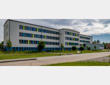 Ein modernes, mehrstöckiges Schulgebäude mit weißer Fassade und farbigen Fensterrahmen in Blau und Grün, umgeben von Grünflächen mit Bäumen. Im Vordergrund befindet sich ein gepflasterter Gehweg und eine Straße.