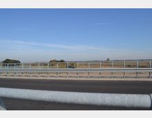 Eine asphaltierte Straße mit metallenen Leitplanken und einem Schutzgitter im Vordergrund, im Hintergrund eine flache Landschaft mit Feldern und Bäumen unter einem klaren blauen Himmel.
