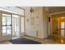 Eingangsbereich eines Gebäudes mit Glastüren, Blick auf eine Außenseite mit Bäumen und parkendem Auto. Innen eine beige Wand mit Informationsplakaten, Wandheizkörper, Uhr und Feuerlöscher; rechts führt ein Korridor zu weiteren Türen.