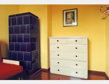 Ein kleiner Raum mit gelben Wänden. Links steht ein dunkelblauer, kachelverkleideter Ofen, daneben eine weiße, sechsschubladige Kommode mit schwarzen Griffen. Über der Kommode hängt ein gerahmtes Porträt eines Kindes.