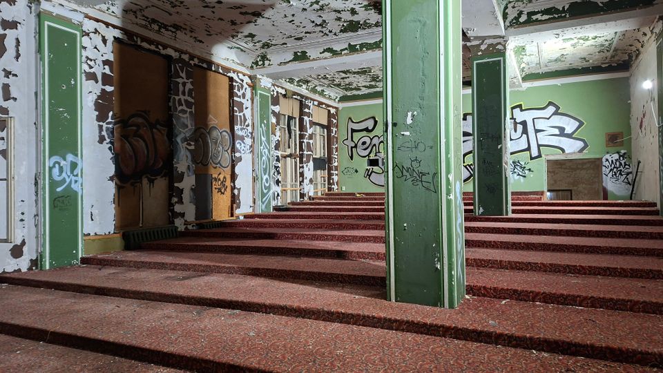 Innenraum eines verlassenen Gebäudes mit breiten, stufenartigen Podesten, grün-weiß gestrichenen Säulen und stark abblätternder Decken- und Wandfarbe. Wände und zugedeckte Fenster sind mit Graffiti bedeckt, im Hintergrund sind alte Holzrahmenfenster und eine kleine Öffnung im Boden zu sehen.