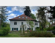 Zweistöckiges weißes Wohnhaus mit rotem Satteldach und mehreren Dachfenstern, umgeben von einem Garten mit Bäumen und Sträuchern. Im Hintergrund ist ein bewölkter Himmel zu sehen.