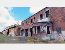 Backsteingebäude in einem verlassenen Industriegebiet mit zerbrochenen Fenstern und verwitterten Fassaden, teils überwuchert von Pflanzen. Im Hintergrund ist eine rostige Lagerhalle mit großen Toren zu sehen, umgeben von einer brachliegenden Fläche.