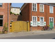 Rote Backsteingebäude mit weißen Fenstern und grünen Fensterläden, flankiert von einem gelben Holztor und einer Reihe geparkter Fahrräder. Der gepflasterte Gehweg und eine ruhige Straße verlaufen im Vordergrund.