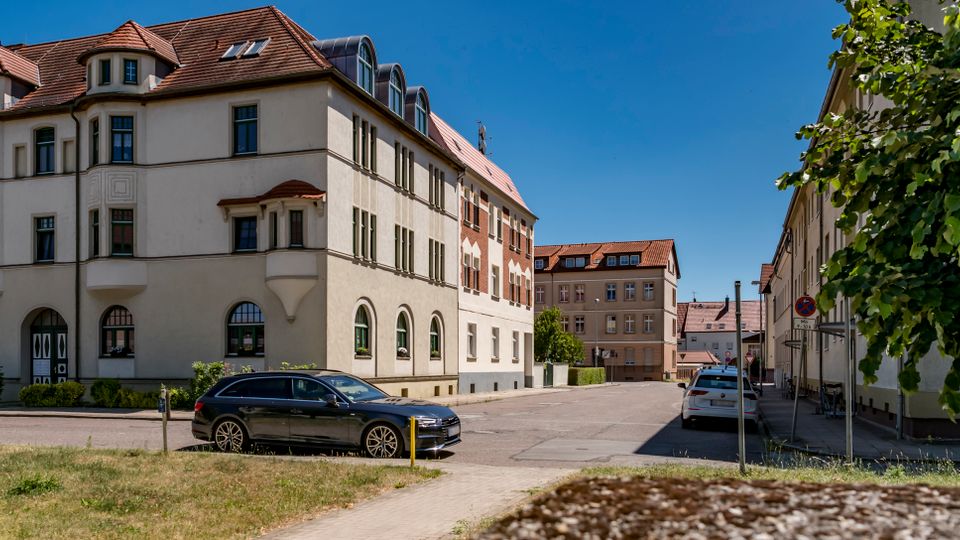 Straßenszene in einem Wohnviertel mit einem mehrstöckigen Altbau mit hellem Putz, Erkern und rotem Ziegeldach an einer Ecke. Rechts verläuft eine ruhige Straße mit weiteren Gebäuden, parkenden Autos und einem Baum am Gehweg unter blauem Himmel.