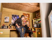 Ein Retro-Zimmer mit Holzdach und grünen Wänden, dekoriert mit Postern, Stickern, und 80er-Jahre Technik wie einem Röhrenfernseher, einem Radio und einer Spielekonsole; zwei Personen sitzen vor dem Fernseher, eine spielt, während der andere zuschaut.