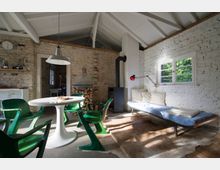 Heller Wohnraum mit sichtbaren weißen Ziegelwänden, einem Esstisch mit grünen Designerstühlen, einem Kaminofen und einer Liege mit blauen Kissen unter einem Fenster. Das Dach aus Holz hat sichtbare Balken, und Holzscheite sind neben dem Kamin gestapelt.