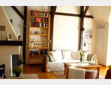 Wohnzimmer mit sichtbaren Holzbalken an der Decke, einem Sofa mit hellen Kissen, einem kleinen Couchtisch aus Holz und Rattanmöbeln. Im Hintergrund steht ein Bücherregal mit bunten Büchern, daneben eine Stehlampe und Wanddekoration.