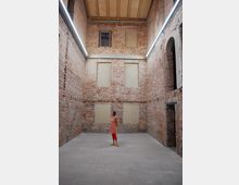 Innenraum eines Backsteingebäudes mit rohen Ziegelwänden, rechteckigen Fensteröffnungen und einem Holzdeckenabschluss. Im Zentrum steht eine Person in bunter Kleidung auf einem schlichten Betonboden.