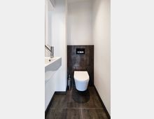 Schmale, moderne Toilette mit wandhängendem WC und dunklen Fliesen an Wand und Boden. Links befindet sich ein weißes Waschbecken mit einem verchromten Wasserhahn.