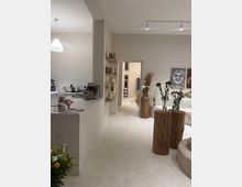 Helles, minimalistisches Innenraumstudio mit weißem Boden und Wänden, links eine Theke mit Kaffeemaschine und Regalen. Rechts stehen Baumstamm-Sockel mit Vasen und Blumenarrangements, daneben Sitzpolster und Wandkunst; ein Durchgang führt in einen hinteren Raum.