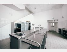 Großküche mit weißen gefliesten Wänden, Edelstahl-Arbeitsflächen, mehreren Kühlschränken und einem Edelstahl-Spülbecken. Der Raum hat eine hohe Decke, gefliesten Boden und eine Doppeltür im Hintergrund.