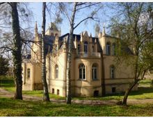 Schlossartiges Gebäude im neugotischen Stil, mit cremefarbener Fassade, spitzen Türmchen und Rundbogenfenstern, umgeben von Bäumen und einer Grasfläche. Im Vordergrund stehen mehrere schlanke Baumstämme.