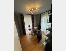 Ein modern eingerichtetes Büro mit einem dunklen Schreibtisch, einem schwarzen Bürostuhl und Accessoires wie einem Lampenschirm und Schreibutensilien. Der Raum hat helle Holzböden, zwei große Fenster mit weißen Vorhängen und dunklen, gemusterten Dekoschals sowie eine Designer-Deckenleuchte.