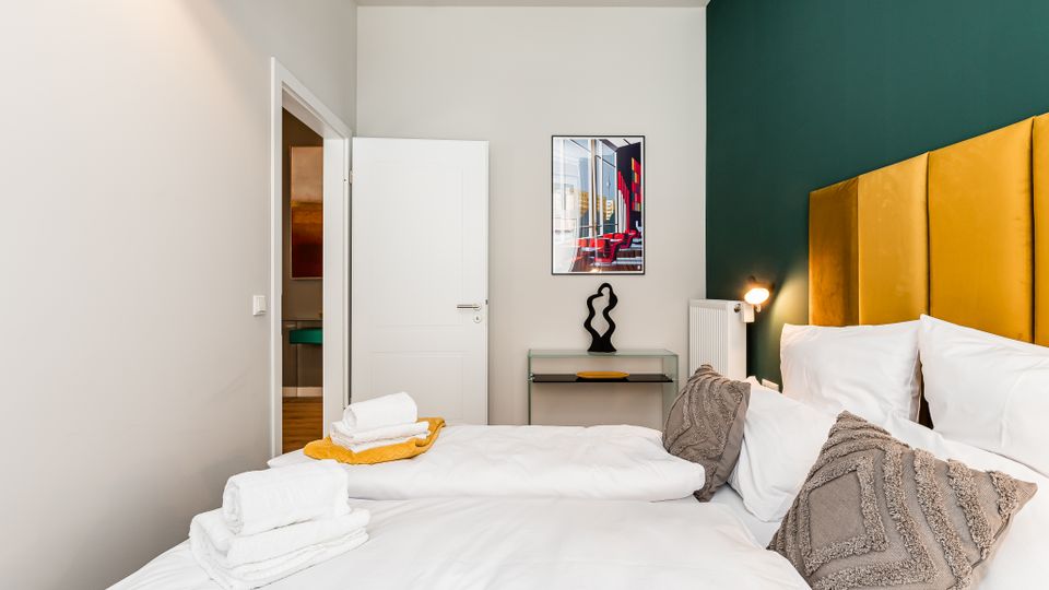 Modernes Schlafzimmer mit Doppelbett, weißer Bettwäsche und senfgelbem, gepolstertem Kopfteil vor einer dunkelgrünen Akzentwand. Neben dem Bett stehen eine kleine Wandlampe, ein Heizkörper und ein Konsolentisch mit Skulptur unter einem gerahmten Bild; links führt eine offene Tür in ein angrenzendes Bad.