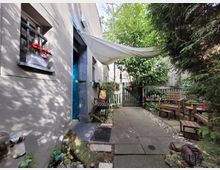 Kleiner Hinterhof mit gepflastertem Weg, umgeben von Pflanzen, einer Sitzgruppe aus Holz und dekorativen Elementen. Links eine Hauswand mit blauer Tür und vergittertem Fenster mit Blumenkasten, im Hintergrund ein kleiner Zaun mit Tor.