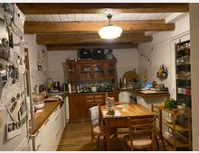 Eine rustikale Küche mit sichtbaren Holzbalken an der Decke, Holzboden und weißen Schränken. In der Mitte steht ein Holztisch mit Stühlen, an der Wand befindet sich ein Holzschrank mit Glasfronten, und links ist ein Kühlschrank mit angehefteten Notizen sichtbar.