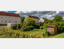 Wohnsiedlung mit hell gestrichenen Mehrfamilienhäusern und roten Ziegeldächern im Hintergrund, umgeben von gepflegten Gärten mit Bäumen, Blumen, Sträuchern, einem Holzschuppen und kleinen Metall- und Plastikhütten. Wolkiger Himmel im Hintergrund.