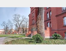 Backstein-Gebäude mit hohen, weißen Fenstern und dekorativen Rundbögen im Vordergrund, umgeben von einem begrünten Park mit Bäumen, Gehwegen und Sträuchern. Links im Hintergrund sind weitere rote Backsteinbauten und ein Torbogen sichtbar.