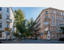 Straßenecke mit gepflasterter Fahrbahn und historischen Wohngebäuden im typisch europäischen Baustil des 19. Jahrhunderts, verziert mit Stuckelementen. Im Vordergrund stehen Verkehrs- und Fußgängerschilder, im Hintergrund Bäume und parkende Autos.