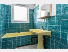 Kleines Badezimmer mit türkisblauen Wandfliesen, Badewanne mit Duscharmatur und einem gelblichen Standwaschbecken. Ein großes Milchglasfenster sowie ein Spiegel mit Ablage befinden sich über dem Waschbecken.