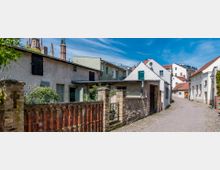 Schmale Kopfsteinpflasterstraße in einem historischen Viertel mit niedrigeren, teilweise verputzten Häusern und alten Ziegelmauern; im Hintergrund sind Kirchtürme und moderne Gebäude sichtbar. Der Himmel ist klar und blau, Bäume und Sträucher säumen den Weg.