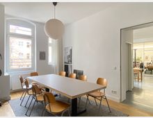 Heller Besprechungs- oder Essraum mit großem rechteckigen Tisch auf Teppich und mehreren Holzstühlen, beleuchtet von einer runden Pendelleuchte. Links große Fenster mit Blick nach außen und Heizkörper darunter; rechts eine breite Öffnung führt in einen angrenzenden Raum mit Werkstatt- oder Büroeinrichtung.