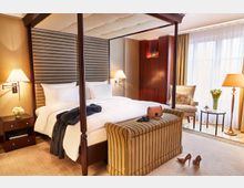 Ein elegantes Hotelzimmer mit einem Himmelbett, flankiert von zwei Nachttischen mit Lampen. Im Vordergrund steht eine gestreifte Polsterbank, im Hintergrund sind ein Sessel, ein Beistelltisch mit Blumen und eine große Fensterfront mit Vorhängen zu sehen.