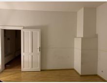 Leerer Innenraum mit hellem Holzboden, weißen Wänden und einer geöffneten weißen Tür, die in einen angrenzenden Raum führt. Rechts an der Wand befindet sich ein hervorstehender, rechteckiger Vorsprung.