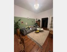 Wohnzimmer mit Holzboden, einer grauen Ecksofa mit bunten Kissen, einem großen cremefarbenen Teppich und einem niedrigen runden Couchtisch. Ein grün gestrichener Wandabschnitt ist mit kleinen Bildern dekoriert, und eine glitzernde Hängelampe befindet sich an der Decke. Links steht ein Holztisch mit Stühlen, und rechts sind weiße Doppeltüren sichtbar.