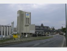 Industriekomplex in städtischer Umgebung mit großen Gebäuden, darunter ein hohes graues Industriegebäude mit der Beschriftung „Asphaltmischwerk“ und angrenzenden Siloanlagen. Im Hintergrund ist ein historisches Lagerhausgebäude mit der Aufschrift „Behala Westhafen“ und ein Kran zu sehen, daneben eine Straße mit Straßenlaternen.