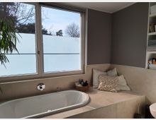 Modernes Badezimmer mit einer weißen Badewanne vor einem Fenster mit halbtransparentem Sichtschutz. Rechts neben der Wanne befindet sich eine geflieste Sitzbank mit Kissen und einer kleinen Korbablage; im Hintergrund ist ein Regal mit Handtüchern und Pflegeprodukten sichtbar.