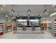 Moderner Ladeninnenraum mit Regalen aus Holz, die Produkte in Verpackungen präsentieren. Im Hintergrund eine große Fotowand mit einer Außenansicht eines Backsteingebäudes und der Begrüßung „Willkommen“ in mehreren Sprachen.