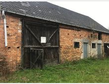 Backsteingebäude mit zerfallener Holzscheune und Metalltüren, umgeben von überwuchertem Gras. Das Dach ist mit dunklen Ziegeln gedeckt, und die Fenster sind mit Gittern versehen.