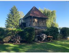 Fachwerkhaus mit schrägem Ziegeldach in einer grünen Gartenlandschaft mit einem Holzzaun, Büschen und einem großen Baum im Hintergrund. Im Vordergrund stehen ein Gartenstuhl, ein Grill und ein kleiner Holztisch auf einer Rasenfläche.