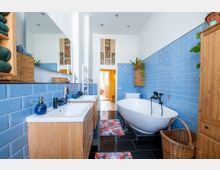 Helles Badezimmer mit blauen, rechteckigen Wandfliesen und schwarzen Bodenfliesen. Der Raum enthält eine freistehende Badewanne, ein Doppelwaschbecken mit Holzmöbeln, dekorative Pflanzen und einen Korb, im Hintergrund ist ein angrenzender Raum mit buntem Fenster erkennbar.