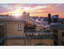 Dachterrasse eines Backsteingebäudes in einer urbanen Umgebung bei Sonnenuntergang, mit Lichterketten dekoriert und einer Gruppe von Menschen, die an Tischen sitzen. Im Hintergrund sind weitere Gebäude und ein farbiger Himmel zu sehen.