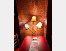 Ein kleiner Tisch mit roter Oberfläche steht vor einer orangefarbenen Wand mit dekorativem, floralen Muster. Drei schräg angeordnete Lampen mit verschiedenen Schirmen beleuchten die Szene; auf dem Tisch liegt eine Getränkekarte und ein runder Untersetzer.