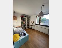 Ein Kinderzimmer mit hellem Holzboden, einem Einzelbett auf der linken Seite und einem Regal mit Büchern und Spielzeug an der Wand. Rechts befindet sich ein Schreibtisch unter einem großen Fenster, das Blick auf einen Garten bietet, und zwei geflochtene Lampenschirme hängen von der Decke.