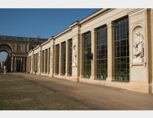 Außenansicht eines historischen Orangeriegebäudes mit großen, rechteckigen Glasfenstern und Sandsteinfassade, ergänzt durch eingebettete Statuen in Nischen. Im Hintergrund ist ein Torbogen mit Säulen zu sehen, flankiert von einer Gartenanlage.