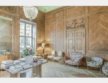Holzvertäfelter Raum im klassischen Stil mit hohen Wänden, großen Fenstern und detaillierten Schnitzereien. Der Raum enthält dekorative Polstermöbel, einen Tisch mit Büchern und Blumenarrangement sowie einen Kronleuchter an einer kunstvoll gestalteten Decke.