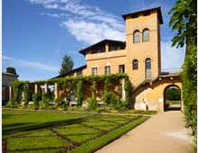 Gelbes Gebäude im mediterranen Stil mit einem Turm und Rundbögen, umgeben von gepflegten Gartenanlagen mit Heckenmustern und Blumenbeeten. Links führt eine überdachte Pergola, während rechts eine Treppe zu einem Torbogen führt.