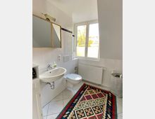 Helles Badezimmer mit weißen Wandfliesen und Bodenfliesen, Waschtisch mit Spiegel, Toilette, Heizkörper unter einem großen Fenster und einem gemusterten Teppich auf dem Boden.