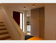 Eingangsbereich eines modernen Hauses mit einer roten Haustür, einem Holztreppenaufgang und Einbauschränken mit weißen Schubladen und Regalböden. Der Boden besteht aus Holz, und an der Decke befinden sich schlichte Deckenspots.