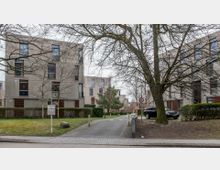 Mehrstöckige Wohngebäude aus Beton mit großen Fenstern, umgeben von Bäumen, Gehwegen und gepflegten Hecken. Im Vordergrund befindet sich eine gepflasterte Zufahrt mit einem Verkehrsschild und einem parkenden Auto.