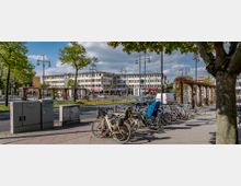 Städtische Straßenszene mit parkenden Fahrrädern im Vordergrund, Bäumen, Laternen und einem begrünten Mittelstreifen. Im Hintergrund stehen mehrstöckige Gebäude mit Geschäften und einem Hotel-Schriftzug.